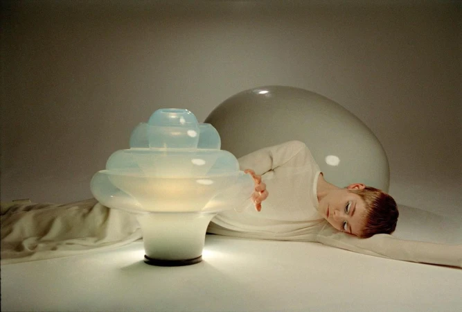 Woman in white next to an illuminated sculptural table lamp and large white sphere in a softly lit interior, modern and minimalist.