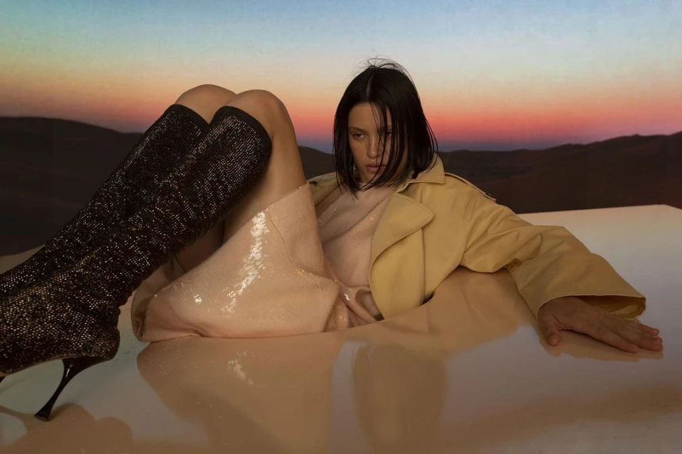 Woman poses in a peach dress and tan trench coat with knee-high black glitter boots against a desert landscape at sunset.