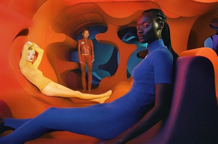 Three fashion models pose in a sculptural orange and blue tunnel wearing blue, red, and yellow outfits. The shot has low lighting and unique colours.