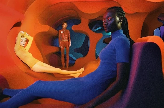 Three fashion models pose in a sculptural orange and blue tunnel wearing blue, red, and yellow outfits. The shot has low lighting and unique colours.