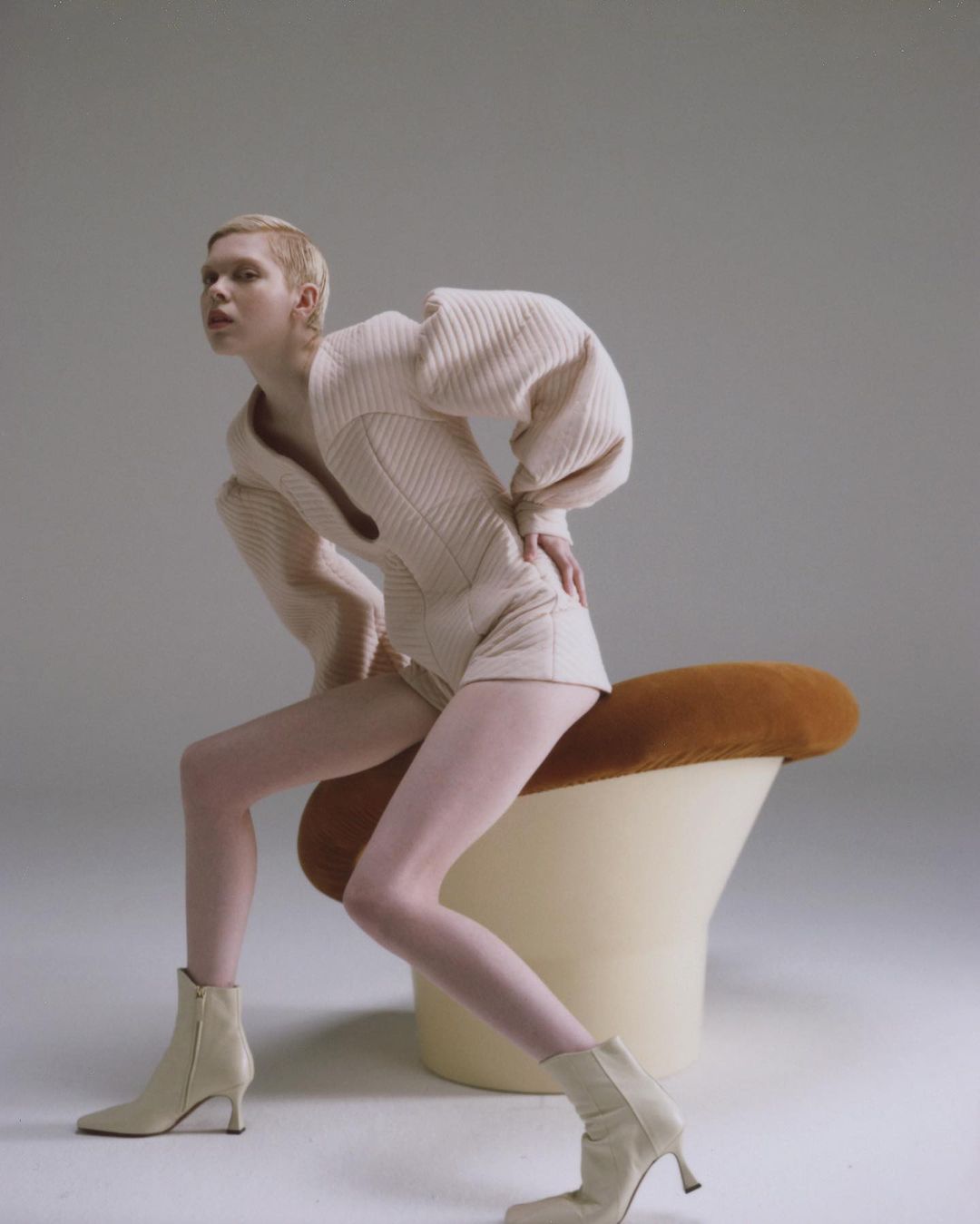 Woman in cream knitwear sits on a mushroom stool against a neutral backdrop in a studio shot. Fashion editorial photography.