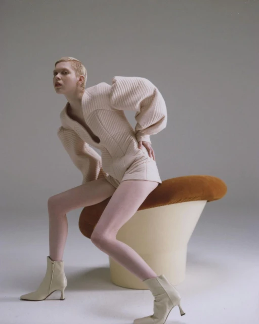 Woman in cream knitwear sits on a mushroom stool against a neutral backdrop in a studio shot. Fashion editorial photography.
