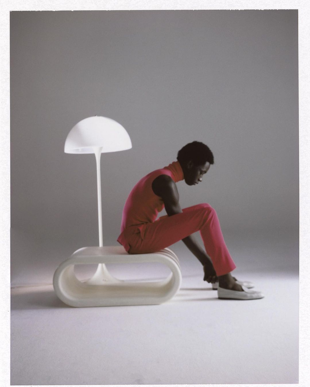 A person in a pink jumpsuit sits on a white, modernist bench next to a white mushroom-shaped lamp in a minimalist photo studio.