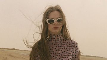 A woman with long brown hair wearing a crocheted turtleneck and white sunglasses stands in front of a neutral-toned desert landscape.