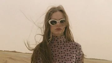 A woman with long brown hair wearing a crocheted turtleneck and white sunglasses stands in front of a neutral-toned desert landscape.