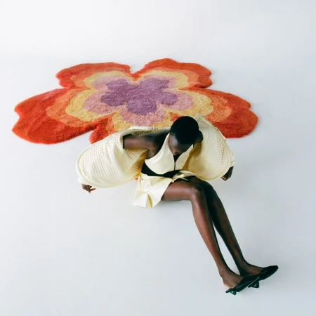 Model wearing a pale yellow dress sitting on white studio floor near a large orange and purple flower-shaped rug