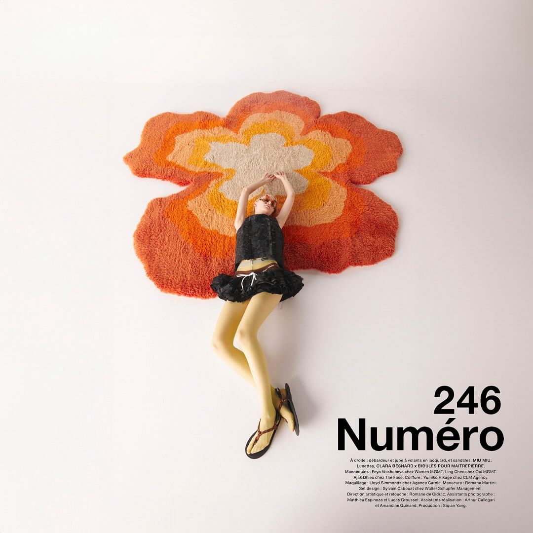 A fair-skinned fashion model lies on a flower-shaped rug in a black top and skirt with white background for magazine ad.