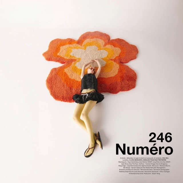 A fair-skinned fashion model lies on a flower-shaped rug in a black top and skirt with white background for magazine ad.