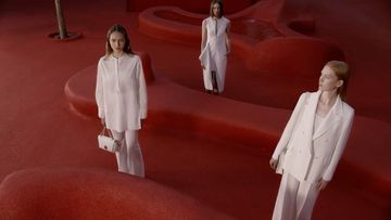 Three women in white outfits stand on a textured red surface, creating a high-fashion editorial image with clean lines and minimalist styling.