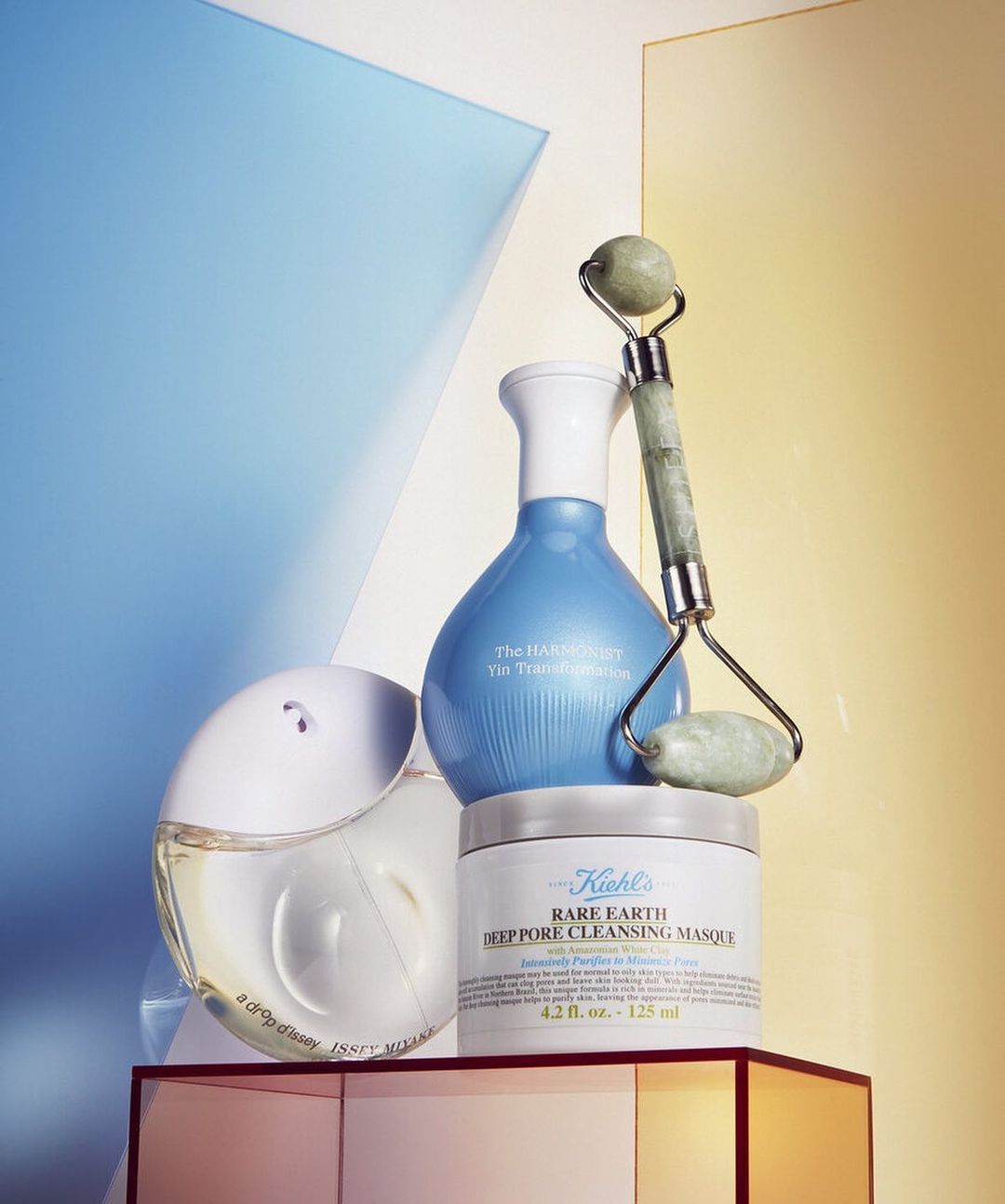 Cosmetic products including a jade roller, a cream, a serum, and a perfume bottle are arranged on a geometric surface with colorful panels.