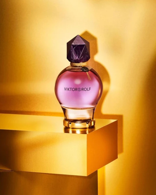 A perfume bottle by Viktor & Rolf sits atop a golden pedestal, illuminated by studio lighting against a rich, golden background.