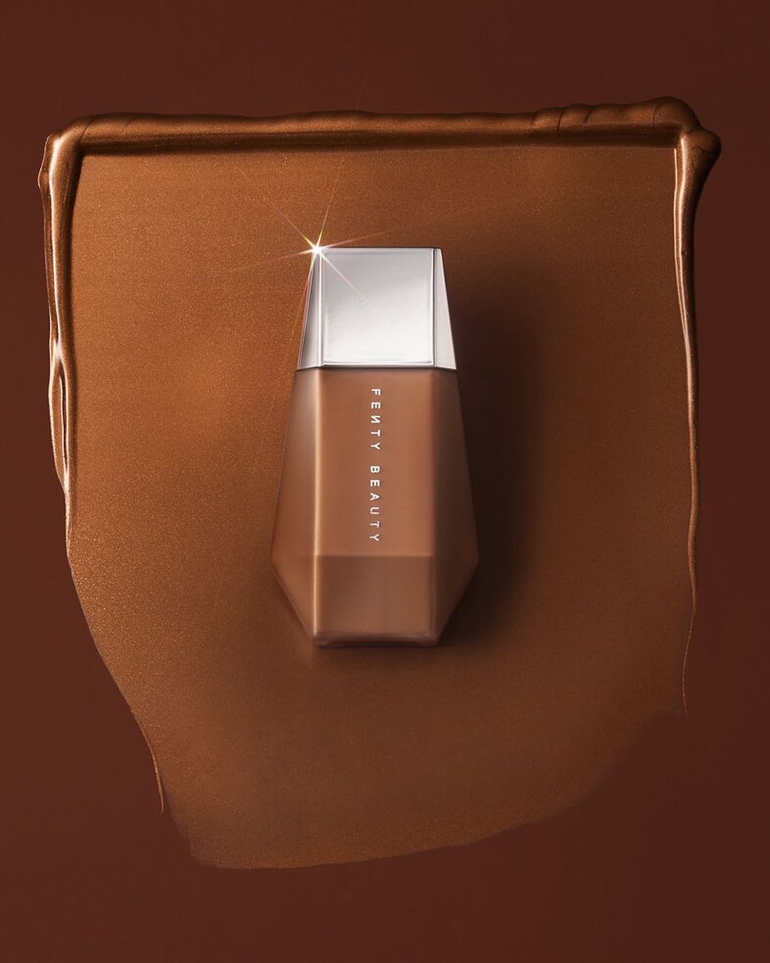 Fenty Beauty foundation bottle in a pool of matching makeup on a brown backdrop, showcasing a beauty product shot.