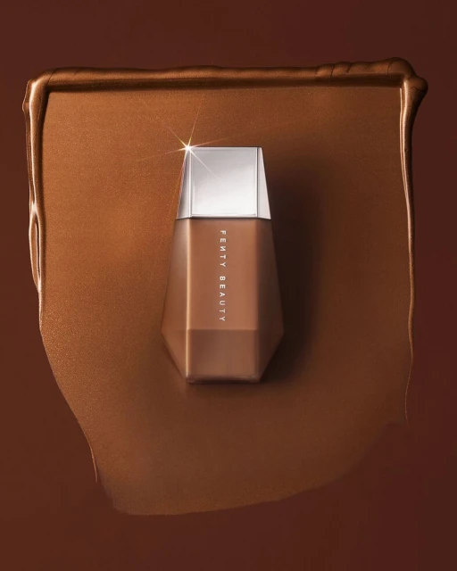 Fenty Beauty foundation bottle in a pool of matching makeup on a brown backdrop, showcasing a beauty product shot.