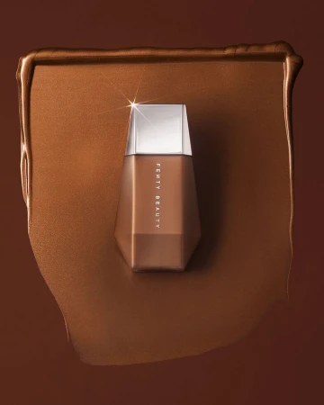 Fenty Beauty foundation bottle in a pool of matching makeup on a brown backdrop, showcasing a beauty product shot.