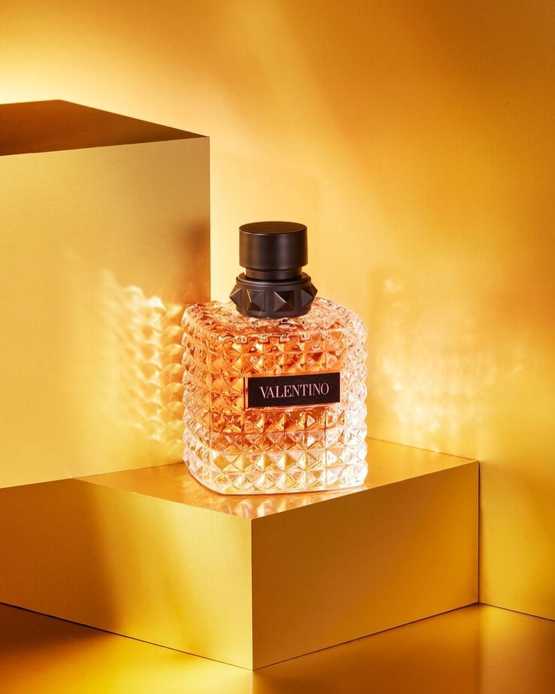 Close-up studio shot of a Valentino perfume bottle with studded design on a geometric gold platform against a matching gold background.