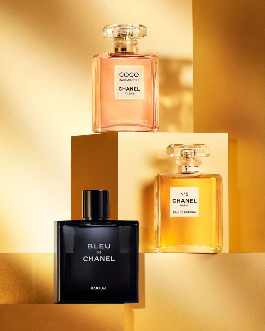 Three Chanel perfume bottles, Coco Mademoiselle, N°5, and Bleu de Chanel, are displayed against a gradient gold background.