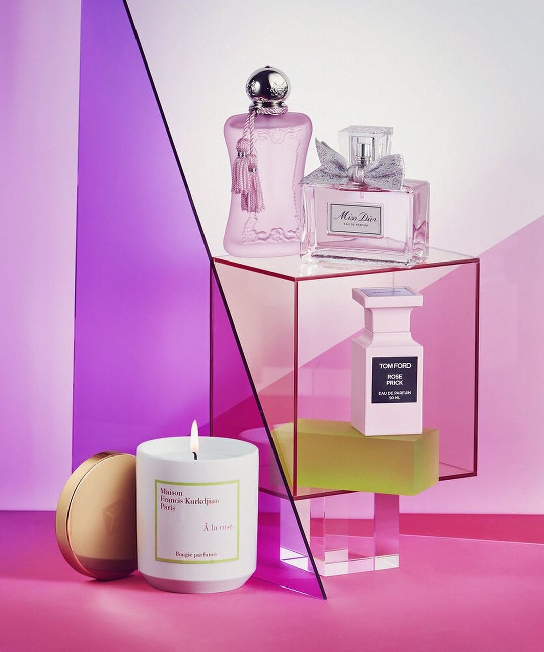 Perfume bottles and candle arranged on transparent blocks with a light purple and pink background for a beauty product display.