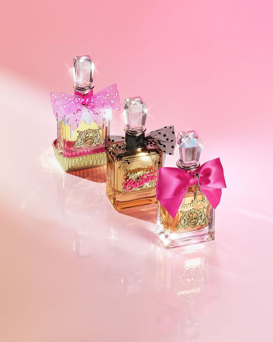 Three Juicy Couture perfume bottles with pink bows are displayed against a gradient pink background, reflecting on a glossy surface.