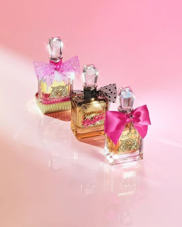 Three Juicy Couture perfume bottles with pink bows are displayed against a gradient pink background, reflecting on a glossy surface.