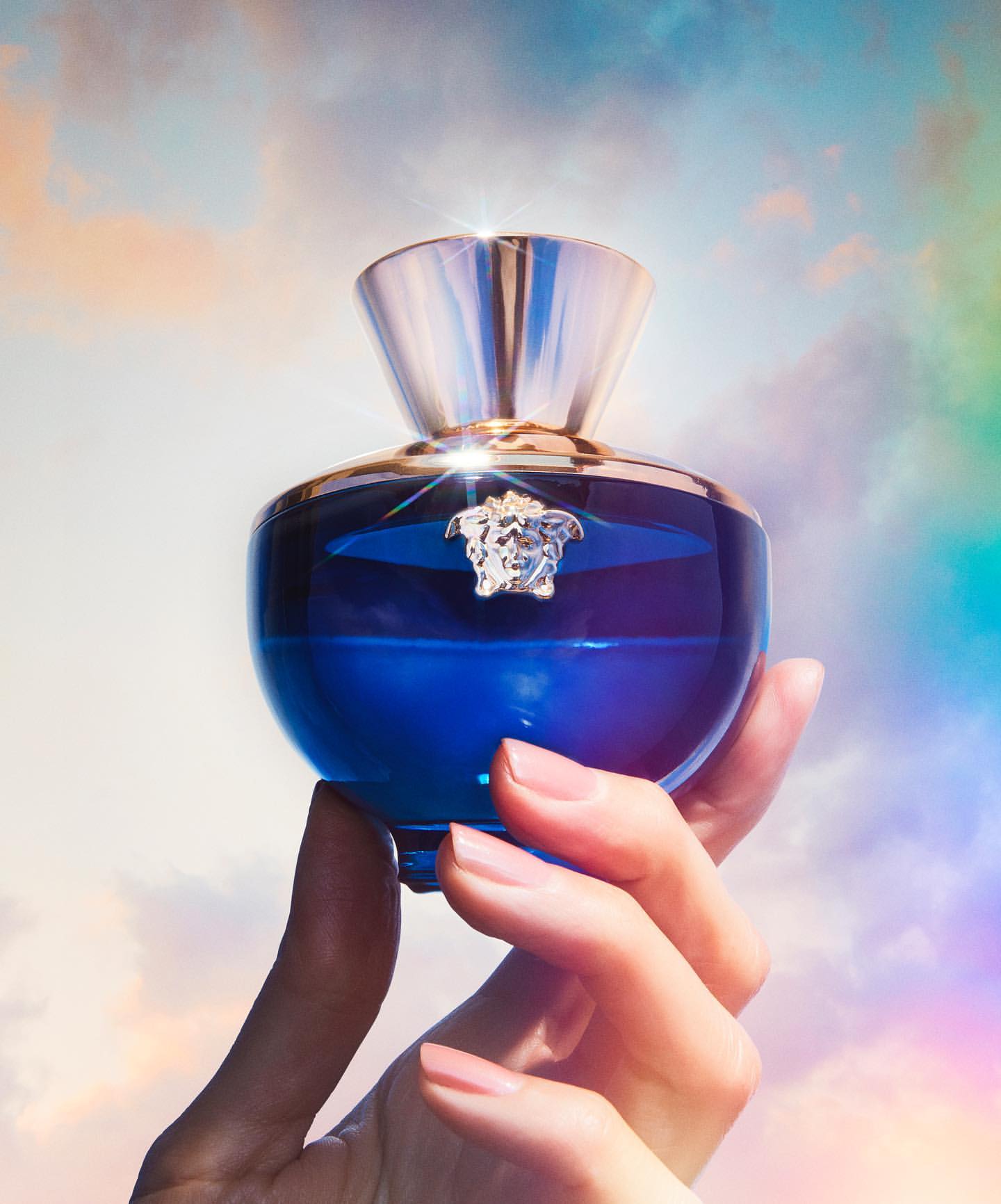 Close-up of a blue perfume bottle held in an adult female's hand against a pastel cloudy sky backdrop, showcasing beauty and elegance.