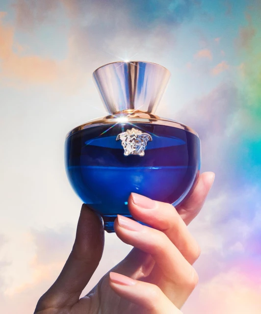 Close-up of a blue perfume bottle held in an adult female's hand against a pastel cloudy sky backdrop, showcasing beauty and elegance.