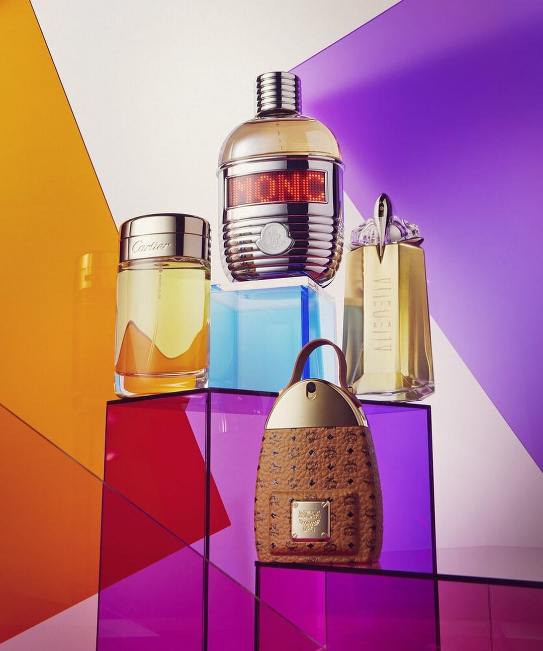 Multiple perfume bottles from brands like Cartier and MCM arranged on colorful transparent geometric platforms in a studio setting.
