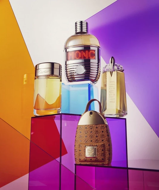 Multiple perfume bottles from brands like Cartier and MCM arranged on colorful transparent geometric platforms in a studio setting.