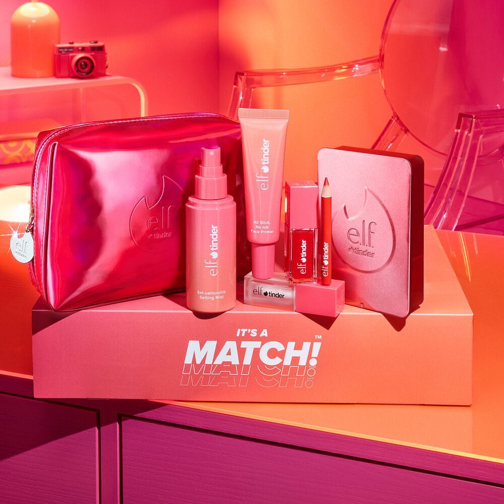 Cosmetics collection by e.l.f. x Tinder featuring makeup bag, spray, primer, lipstick, pencil, and compact on display in a studio setting.