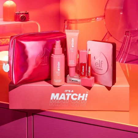 Cosmetics collection by e.l.f. x Tinder featuring makeup bag, spray, primer, lipstick, pencil, and compact on display in a studio setting.