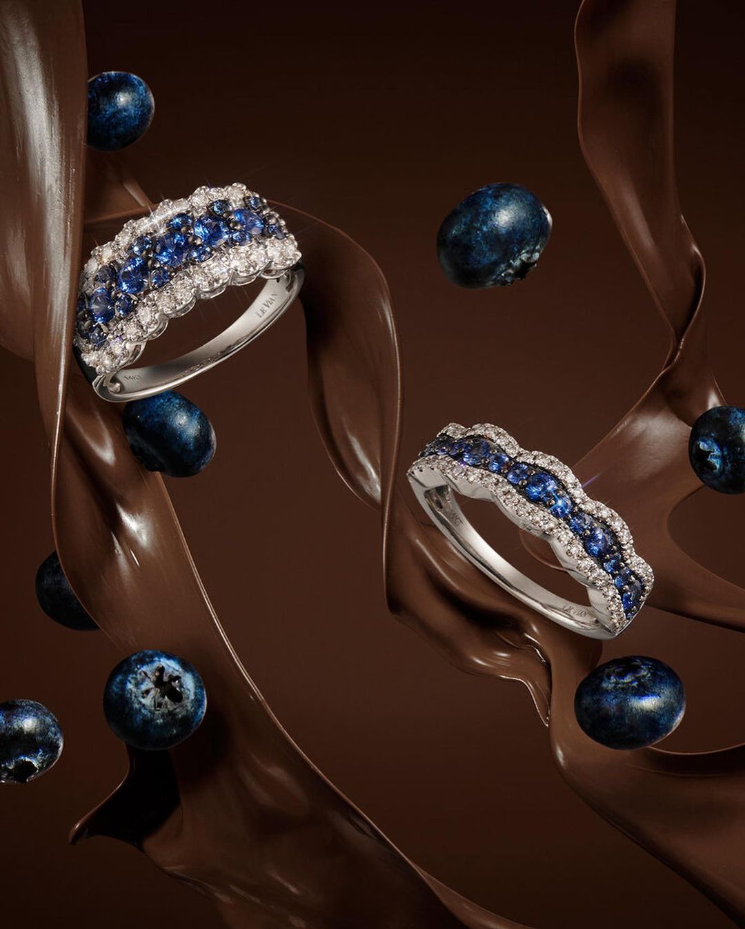 Jewellery product shot featuring diamond and blue sapphire rings, fresh blueberries, and chocolate sauce against a dark brown background.