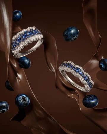 Jewellery product shot featuring diamond and blue sapphire rings, fresh blueberries, and chocolate sauce against a dark brown background.