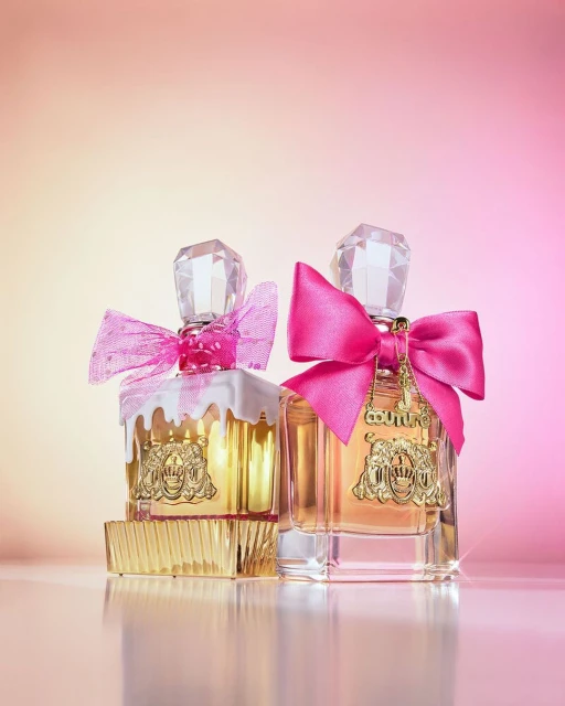 Two Juicy Couture perfume bottles with pink bows sit side-by-side on a glossy surface against a gradient pink background in a beauty shot.