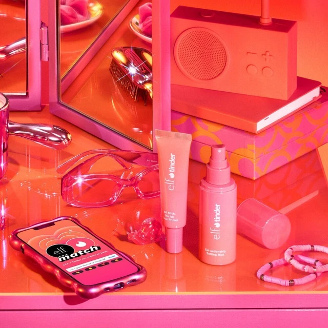 Still life of e.l.f. and Tinder makeup products in pink and coral tones, including a primer, mist, phone case, and accessories.
