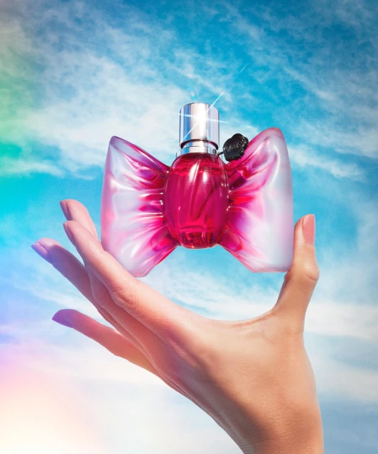 A hand holds a perfume bottle with a pink bow against a blue sky with clouds, creating a vibrant and eye-catching beauty product shot.