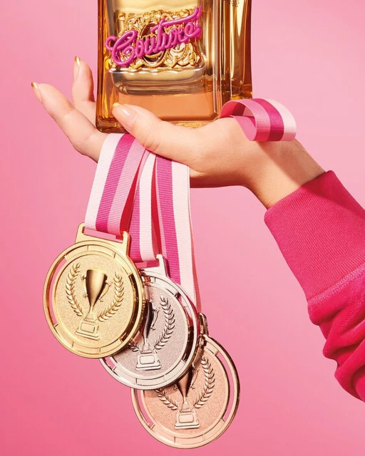 Juicy Couture perfume bottle held in a woman's hand, displayed against a pink background with three hanging gold medals.