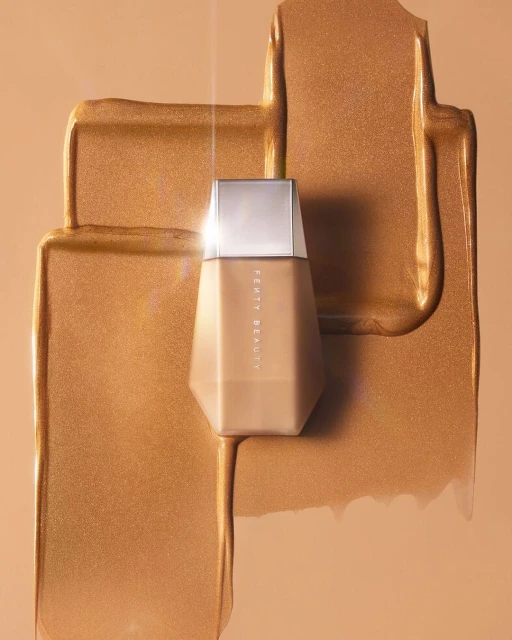 Fenty Beauty foundation bottle with smears of matching makeup on a neutral tan background. Silver lid on an octagonal bottle.