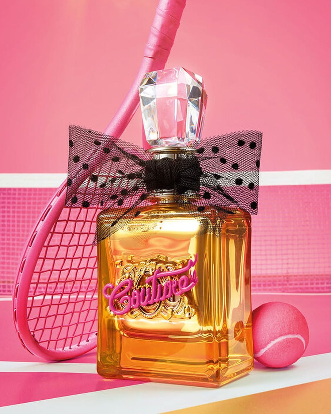 Juicy Couture perfume bottle with black bow, pink tennis racket, and pink ball on a pink and white tennis court.