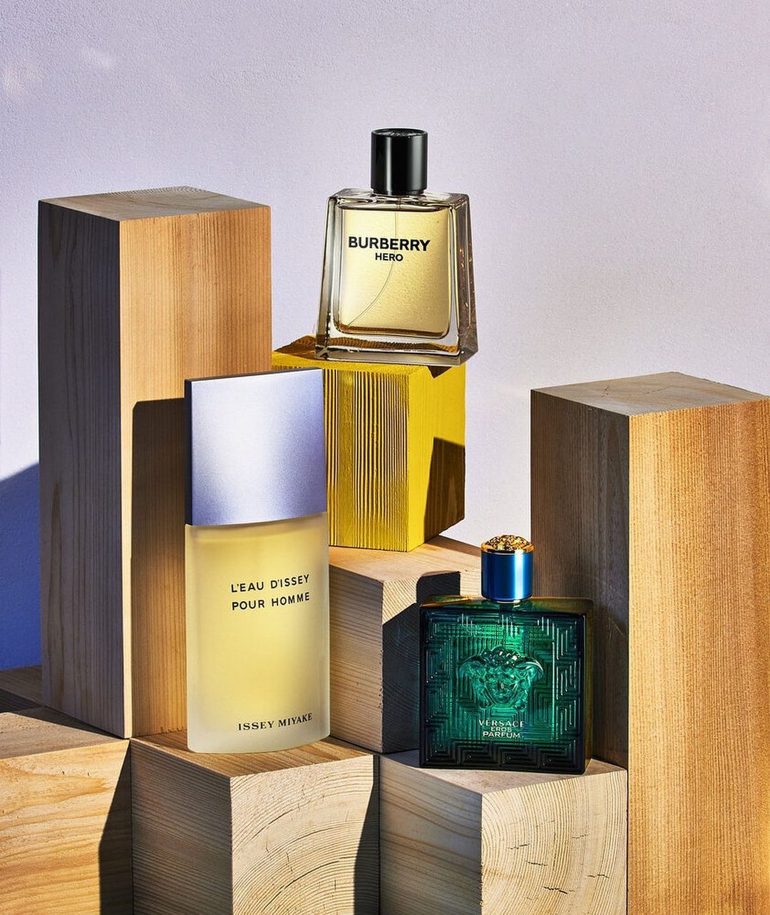 Three luxury men's fragrance bottles, Burberry Hero, Issey Miyake L'Eau D'Issey Pour Homme, and Versace Eros Parfum are stacked on wooden blocks.
