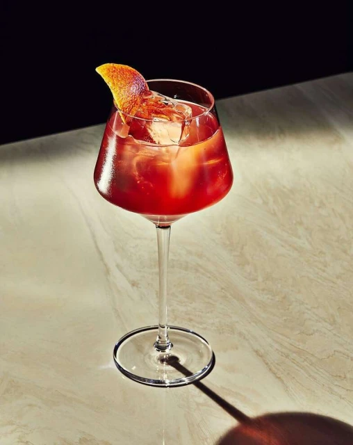 A stemmed glass contains a red cocktail with ice and an orange peel garnish, set on a marble surface against a dark background.