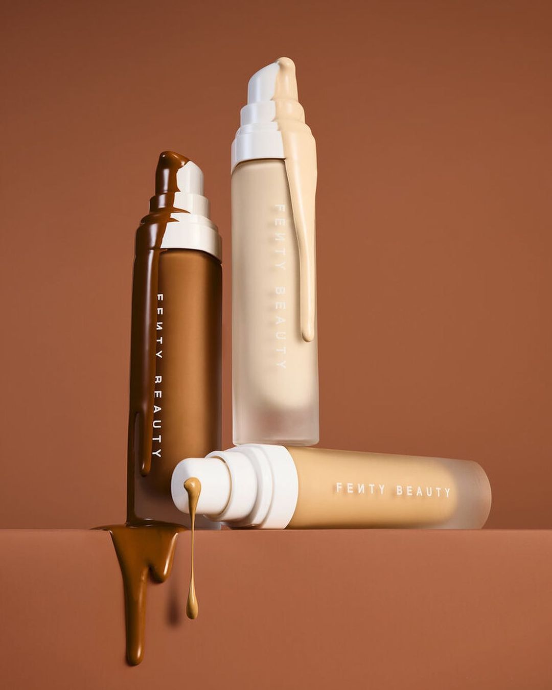 Three bottles of Fenty Beauty foundation in different shades, with foundation dripping, set against a brown background. Beauty product shot.