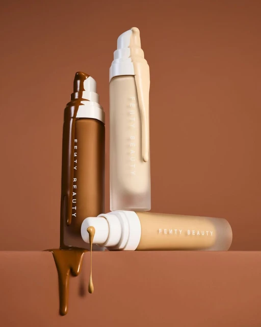 Three bottles of Fenty Beauty foundation in different shades, with foundation dripping, set against a brown background. Beauty product shot.