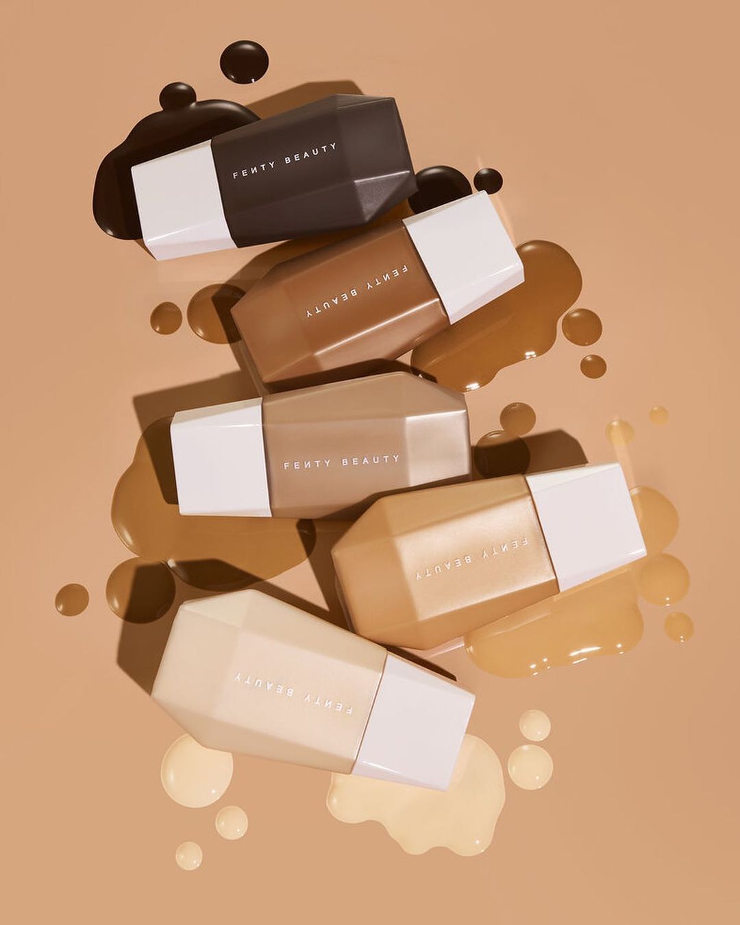 Five Fenty Beauty foundation bottles in varying shades are stacked together on a tan surface with matching foundation splatters.