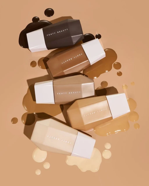 Five Fenty Beauty foundation bottles in varying shades are stacked together on a tan surface with matching foundation splatters.