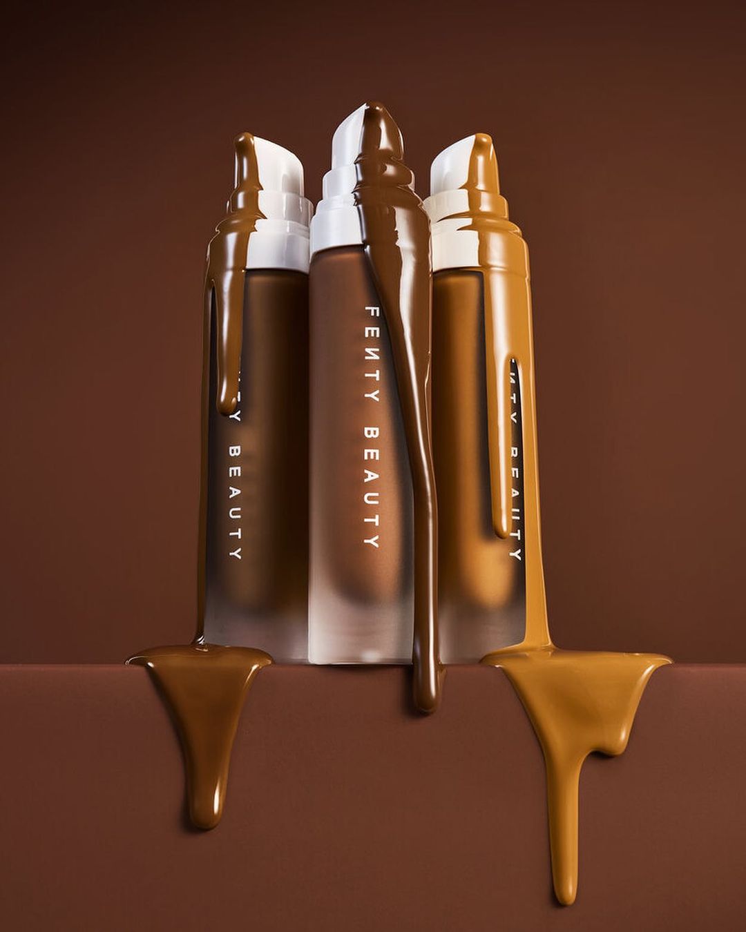 Three bottles of Fenty Beauty foundation in various shades stand upright, each bottle partially covered with matching liquid foundation.