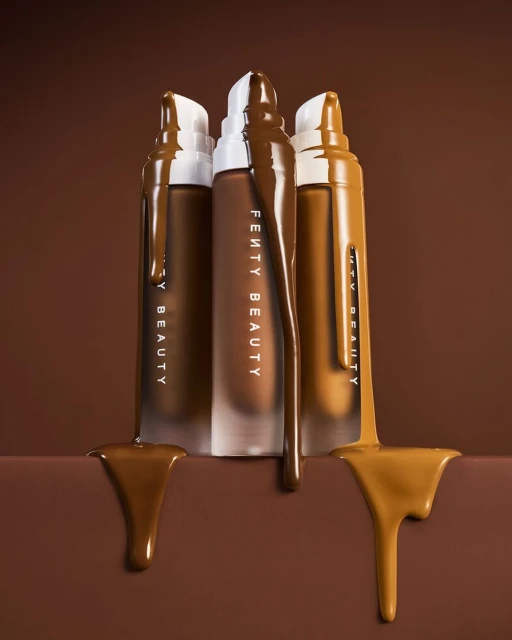 Three bottles of Fenty Beauty foundation in various shades stand upright, each bottle partially covered with matching liquid foundation.