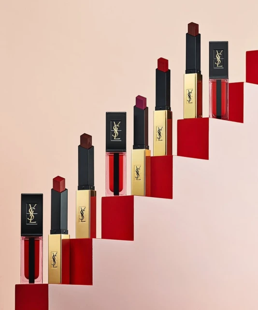 Collection of YSL lipsticks and lip glosses in red, black, and gold packaging arranged on stacked red and light pink steps.