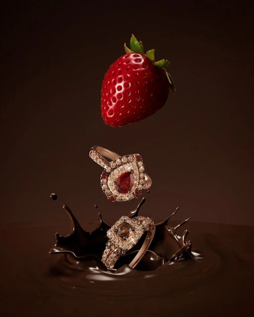 Elegant jewelry advertisement featuring two rings with gemstones, a fresh strawberry, and a splash of rich, dark chocolate against a brown backdrop.