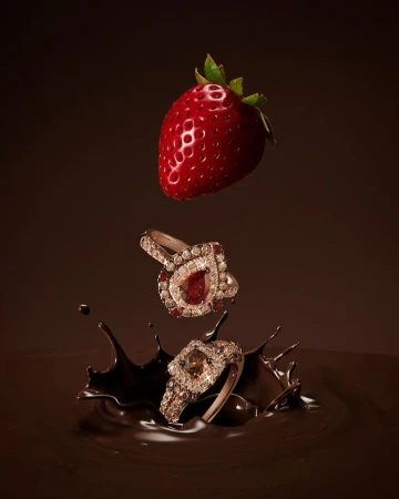 Elegant jewelry advertisement featuring two rings with gemstones, a fresh strawberry, and a splash of rich, dark chocolate against a brown backdrop.