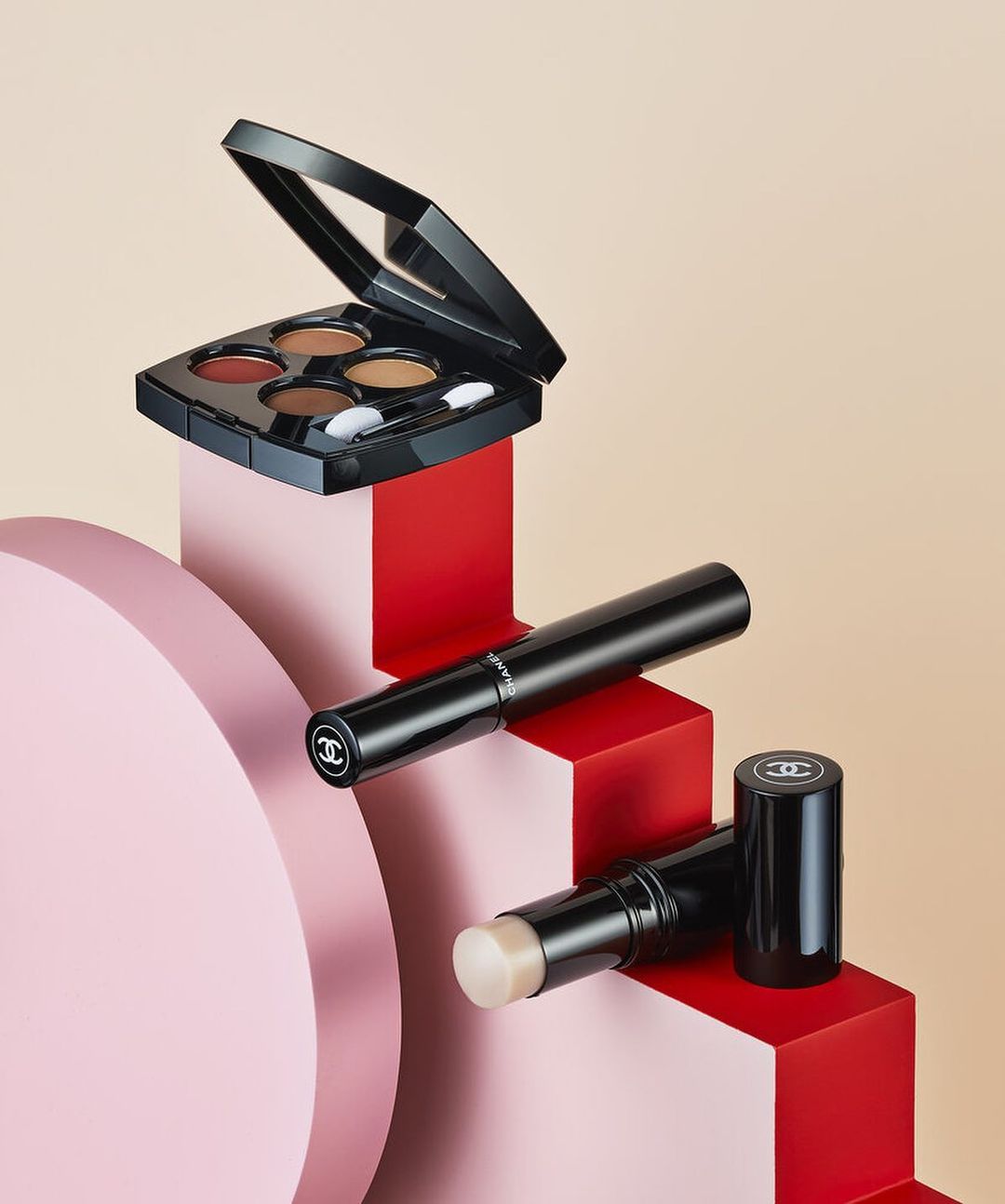 High-angle product shot of Chanel makeup: an eye shadow palette, a mascara tube, and a face stick, stacked on pink and red geometric blocks.