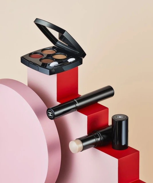 High-angle product shot of Chanel makeup: an eye shadow palette, a mascara tube, and a face stick, stacked on pink and red geometric blocks.
